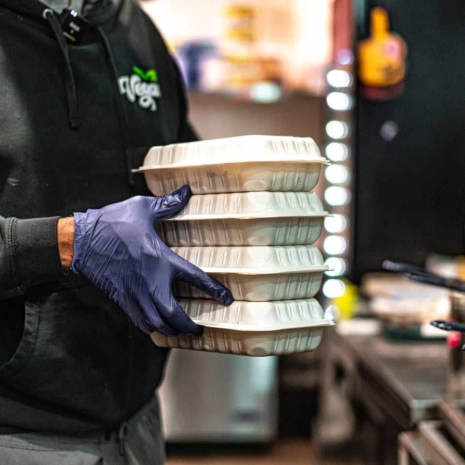 A person wearing a Vegan On The Fly hoodie and blue gloves carries a stack of takeout containers.