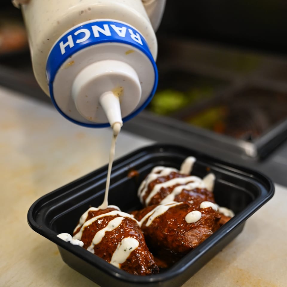 Vegan wings drizzled with ranch dressing from a squeeze bottle, in a black takeout container.