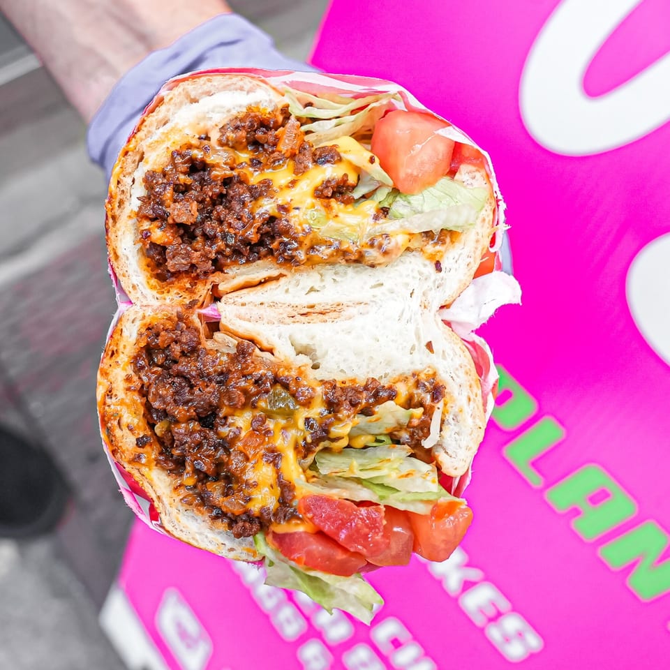 A vegan chopped cheese sandwich, cut in half, with lettuce and tomato, held over a pink surface.