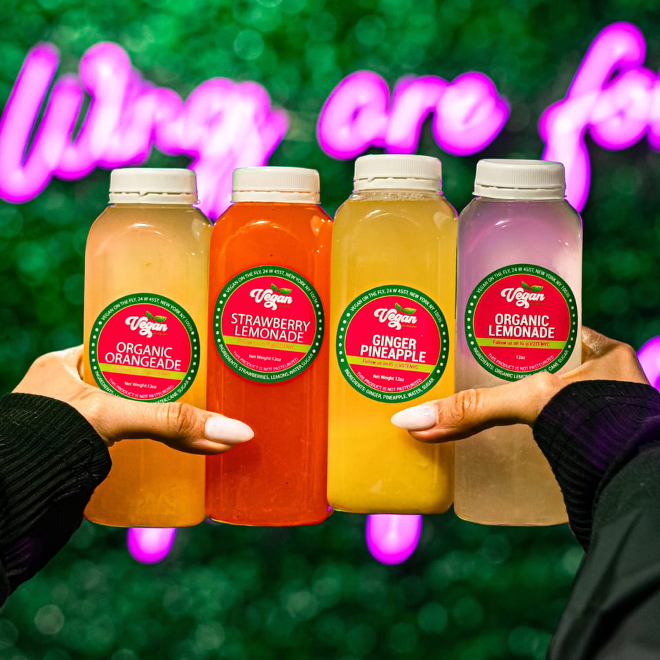 Hands holding four Vegan On The Fly juice bottles: orangeade, strawberry lemonade, ginger pineapple, and lemonade.