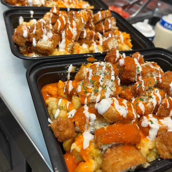 Vegan mac and cheese topped with buffalo 'chicken' and ranch dressing in black takeout containers.