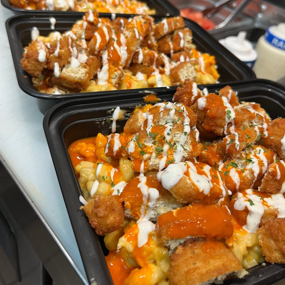 Vegan mac and cheese topped with buffalo 'chicken' and ranch dressing in black takeout containers.