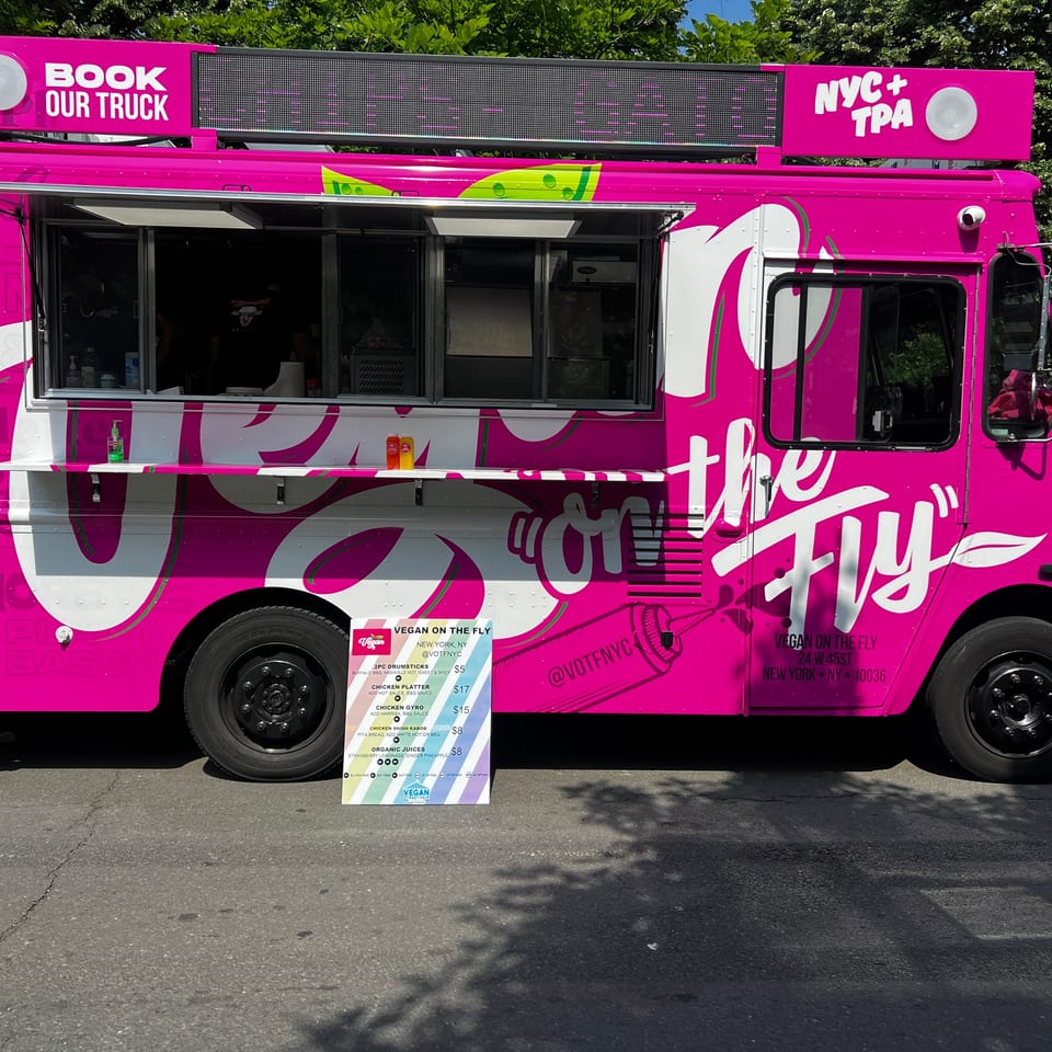 A vibrant pink Vegan On The Fly food truck with a menu board parked on a street.