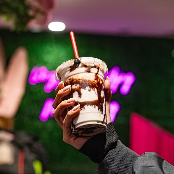 Hand holding a vegan milkshake with chocolate drizzle and a red straw against a neon background.