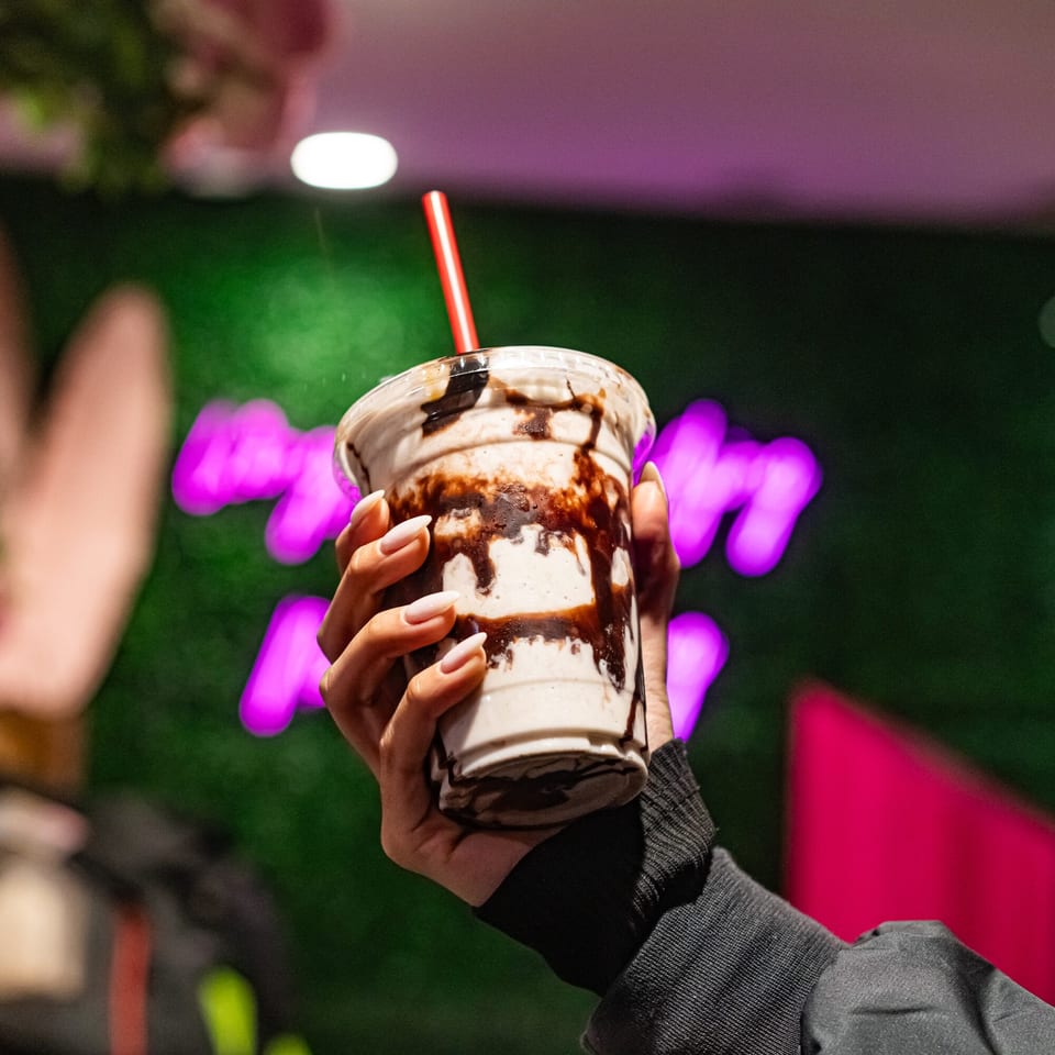 Hand holding a vegan milkshake with chocolate drizzle and a red straw against a neon background.