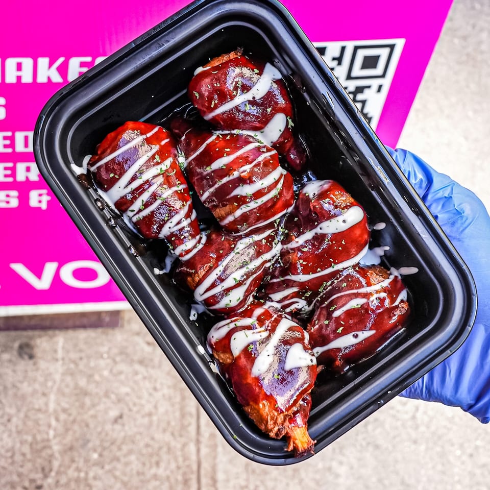 A hand in a blue glove holds a black container of vegan wings drizzled with white sauce and herbs.