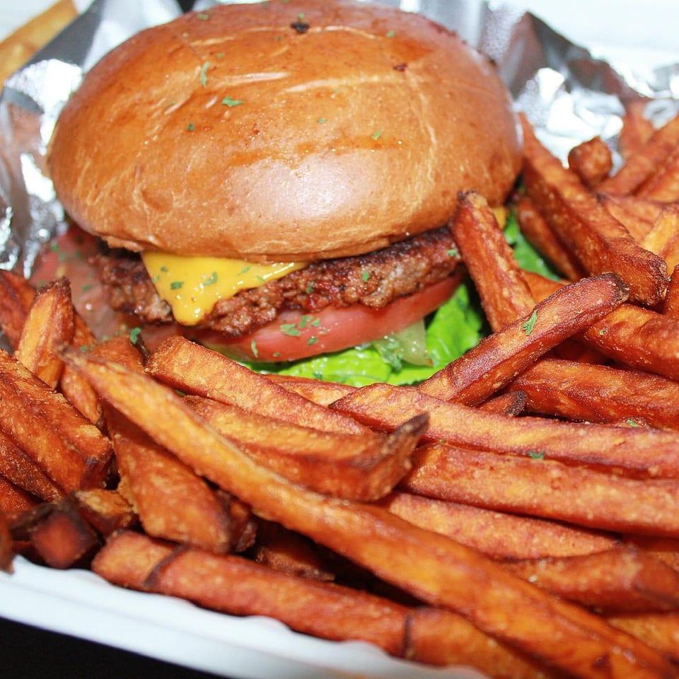Vegan burger with melted cheese, lettuce, and tomato, served with sweet potato fries.