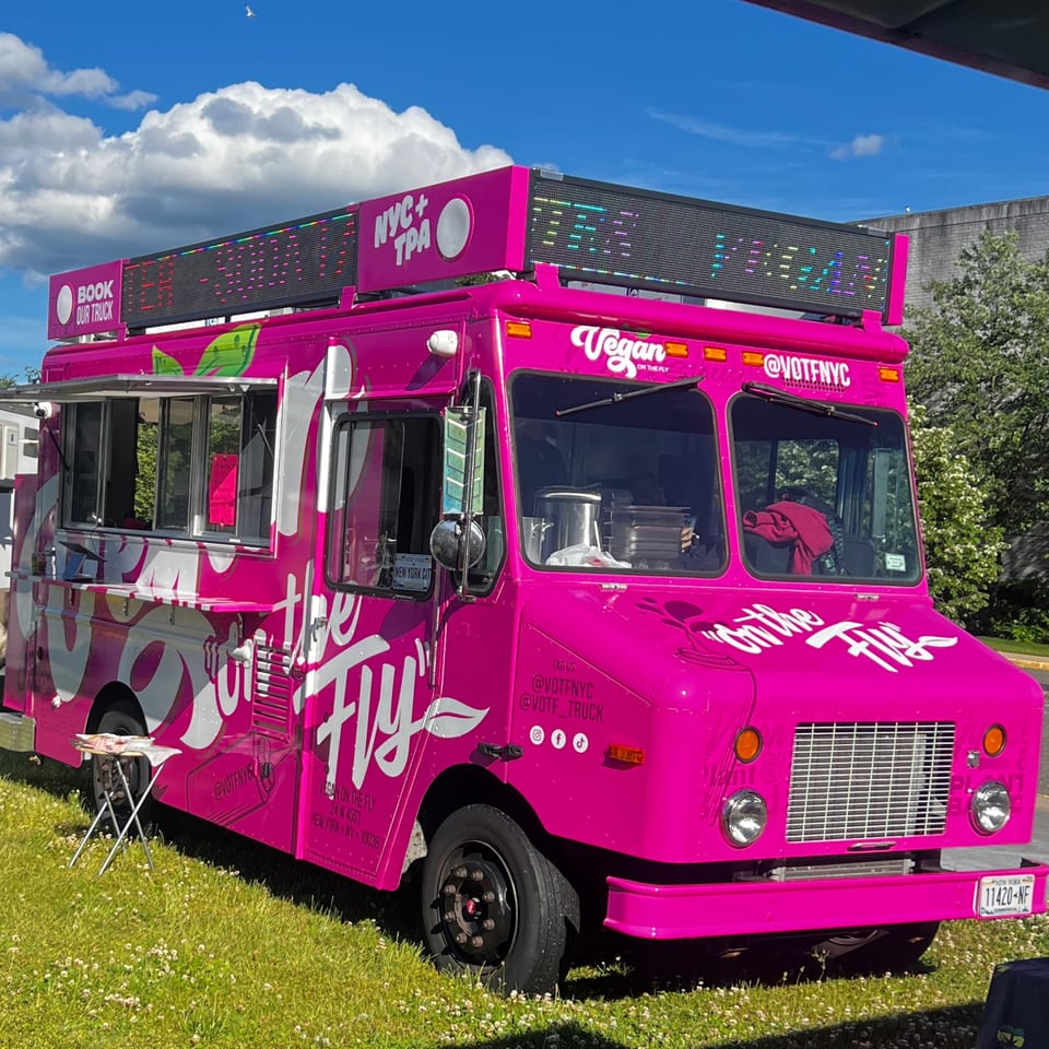 Bright pink Vegan On The Fly food truck with LED signs, parked on green grass under a blue sky.